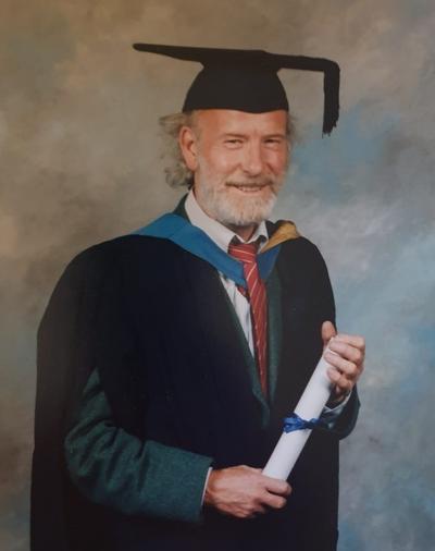 Thomas wears black graduation cap and cape whilst holding white scroll with blue ribbon as he smiled for a graduation day photo. 