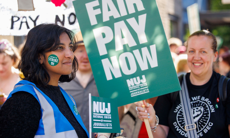 Woman with dark hair and blue and silver hi-vis jacket is pictured with green sticker on cheek in front of a person holding fair pay now placard 