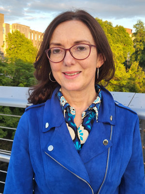 Máire T Ní Mhadaoin in blue jacket and dark spectacles smiles for a photo. 