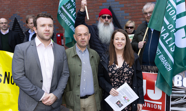 Journalist Barry McCaffrey is photographed next to a man to his left and woman to his right outside protect. NUJ green flags are behind them.