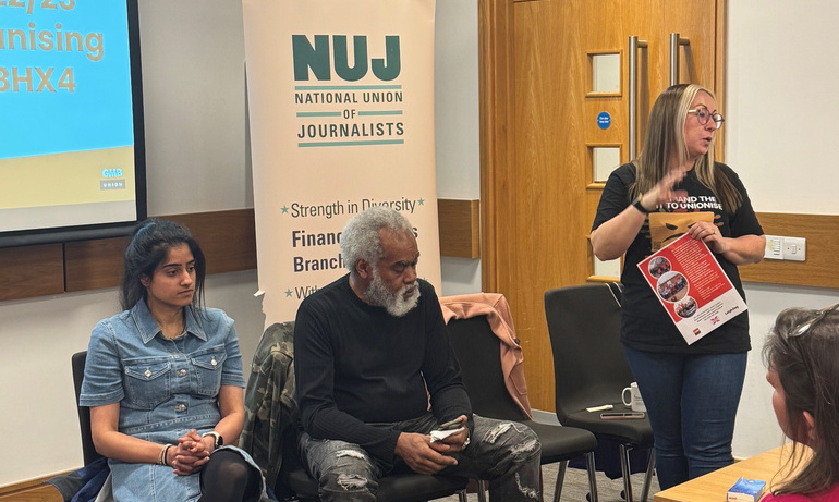 Woman delivers presentation to audience, white roller banner with NUJ logo is visible, half of Powerpoint presentation can be seen.