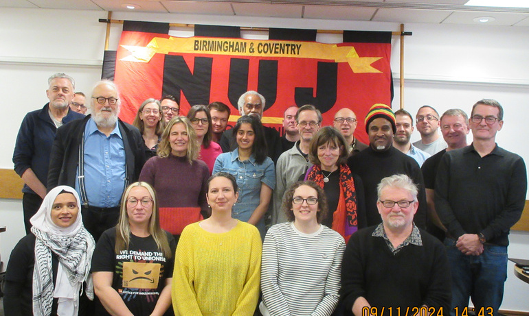 Reps pose for a photo smiling, large NUJ flag visible hanging on wall in red and yellow with black writing. 