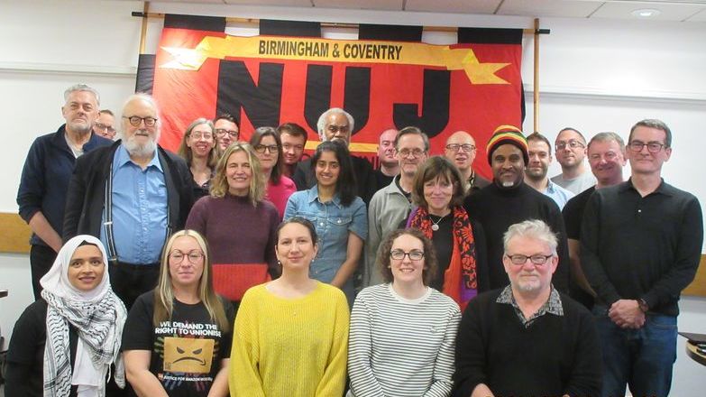 Reps pose for a photo smiling, large NUJ flag visible hanging on wall in red and yellow with black writing. 