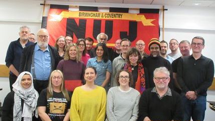 Reps pose for a photo smiling, large NUJ flag visible hanging on wall in red and yellow with black writing. 