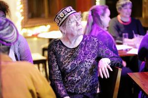 Ann is pictured in a dark patterned blouse wearing glasses and a hat. She is seated. People are sitting at tables in the background.