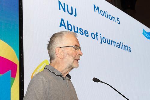 Anton McCabe wearing spectacles, grey shirt with black and white collar trim speaks into mic with slide visible in background - text on slide says motion 5 NUJ Abuse of journalists