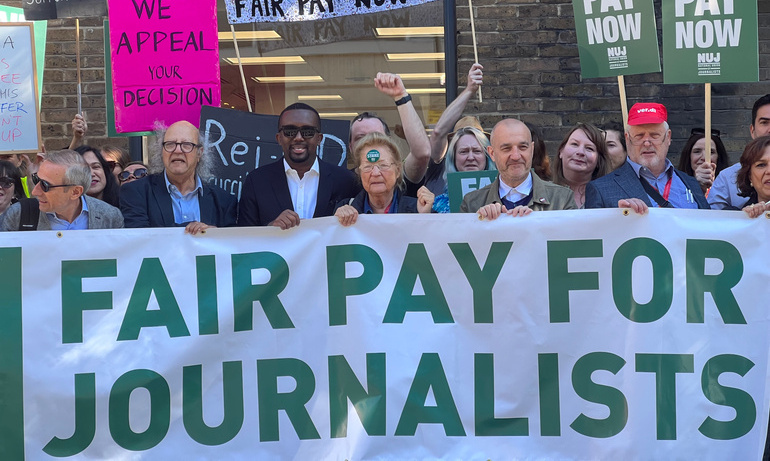 Large white and green fair pay for journalists banner is held by men and women, placards with fair pay now in white on green background visible.