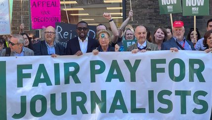 Large white and green fair pay for journalists banner is held by men and women, placards with fair pay now in white on green background visible.