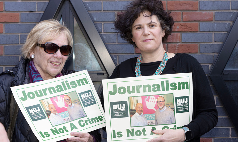 Claire Hanna poses with another who wears dark sunglasses, they both hold posters with text 'journalism is not a crime' and image of journalists.