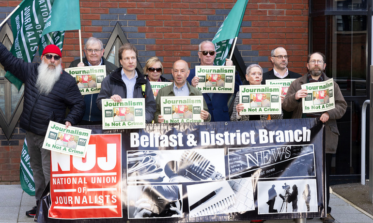 Attendees hold posters and wave NUJ flags as they pose for a photo with NUJ Belfast& District branch banner with red NUJ logo.