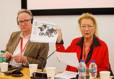 IFJ Presient Dominique Pradalie in red jumper and black spectacles holds document with map of journalists killed. Sits next to Tim Dawson in brown blazer, black spectacles and wears black headset. 