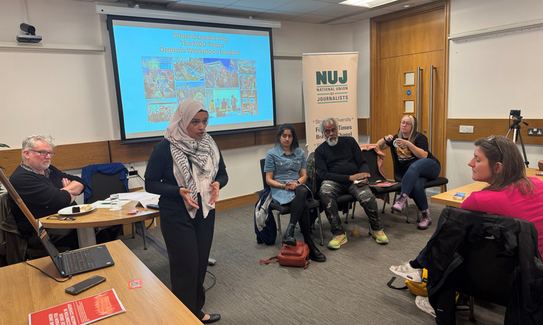 Woman delivers presentation to audience, half of whom sit at desks. Powerpoint presentation shown in background, NUJ roller banner visible.